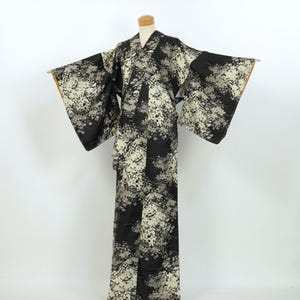 May include: A full-length kimono featuring a black base color with an all-over pattern of cream and light gray floral clusters. The garment has wide, extended sleeves and a wrap-front design, displayed on a mannequin against a plain background.