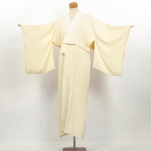 May include: A cream-coloured kimono displayed on a mannequin. The kimono features long, wide sleeves and a wrap-style front. The fabric has a subtle, textured pattern. The collar is white. The garment is displayed on a metal stand.