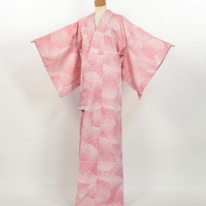 May include: A pink and white patterned kimono displayed on a mannequin. The kimono features a V-neck and wide sleeves. The fabric has a repeating pattern of white shapes on a pink background. The garment is designed for a relaxed fit.