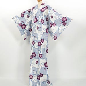 Puede incluir: Un yukata blanco con un estampado floral azul y morado. El yukata tiene un diseño japonés tradicional con un cuello ancho y mangas largas.
