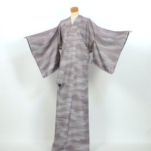 May include: A full-length kimono in a muted mauve-gray tone featuring a soft, horizontal blurred or streaked pattern throughout the fabric. The garment has wide, extended sleeves and a structured collar, displayed on a mannequin form.