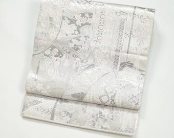 May include: A folded, ornate kimono in shades of white and silver. The fabric features a detailed pattern of floral and geometric designs. The kimono is folded neatly, showcasing the intricate details of the fabric.