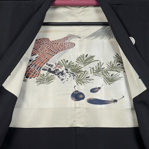 Könnte beinhalten: Schwarzer Kimono mit cremefarbenem Futter, das eine detaillierte Illustration zeigt. Die Illustration zeigt einen braunen und weißen Adler, grüne Kiefernzweige und blaue und schwarze Auberginen. Der Kimono hängt an einem schwarzen Kleiderbügel.