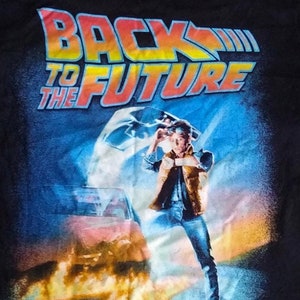 Könnte beinhalten: Ein schwarzes T-Shirt mit einem Grafikdruck des Films "Zurück in die Zukunft". Die Grafik zeigt einen DeLorean, eine Zeitmaschine, mit Flammen, die aus dem Heck kommen, und Marty McFly, der vor dem Auto steht. Der Text "Zurück in die Zukunft" ist in Gelb, Orange und Rot gedruckt.