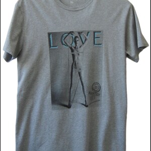May include: A grey t-shirt with a graphic print of a magazine cover featuring the word "LOVE" in large blue letters and a photo of a person standing in a circle. The magazine cover also includes the text "KRISTEN" and a logo.