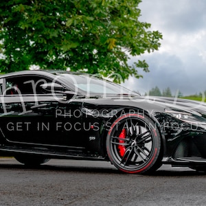 May include: A black Aston Martin sports car with red brake calipers and a license plate that reads "007D". The car is parked on a paved road with a green grassy area in the background.