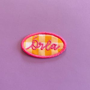 May include: An oval-shaped embroidered patch featuring a yellow and white gingham check pattern. The name Orla is stitched across the centre in a bright pink cursive font, and the entire edge is finished with a thick, bright pink stitched border.