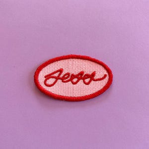 May include: An oval-shaped embroidered patch featuring a light pink background with a bright red stitched border. The name Jess is embroidered in the centre in a cursive red script font, creating a clear contrast against the light pink fabric.