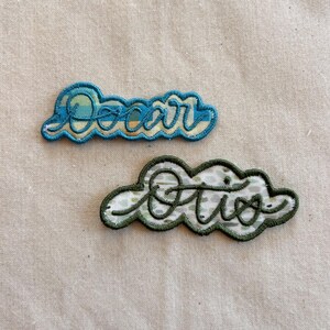 May include: Two embroidered fabric patches with scalloped edges. The top patch features the name Oscar in blue cursive thread over a blue and yellow striped background. The bottom patch features the name Otis in green thread over a green patterned background.
