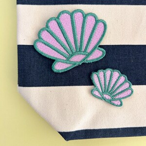 May include: Two embroidered seashell patches featuring light pink centres and dark green stitched borders. The patches are displayed on a fabric surface with horizontal navy blue and cream-coloured stripes, creating a nautical-inspired aesthetic.