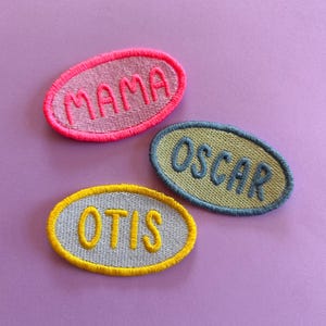 May include: Three oval-shaped embroidered patches with stitched borders. The top patch is light pink with hot pink text reading MAMA. The middle patch is olive green with dark blue text reading OSCAR. The bottom patch is light grey with yellow text reading OTIS.