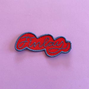 May include: A red embroidered patch with a blue border featuring the name Finlay written in a blue cursive script. The patch has a wavy, cloud-like shape and is designed for attachment to fabric items through sewing or heat application.