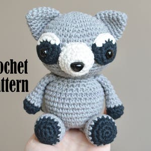 May include: Crochet pattern for a gray amigurumi raccoon with black eyes and a white nose. The raccoon is sitting on a hand.
