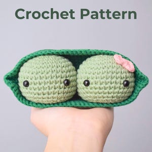 May include: Two crocheted peas in a green pod with black eyes. The pea on the right has a pink flower on top.