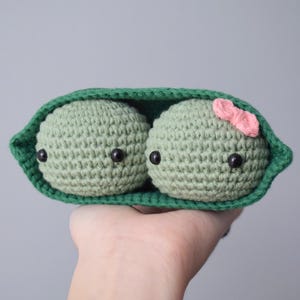 Pu&ograve; includere: Due piselli all'uncinetto in un baccello verde, uno con un fiocco rosa. I piselli hanno occhi neri e sono fatti con filato verde.