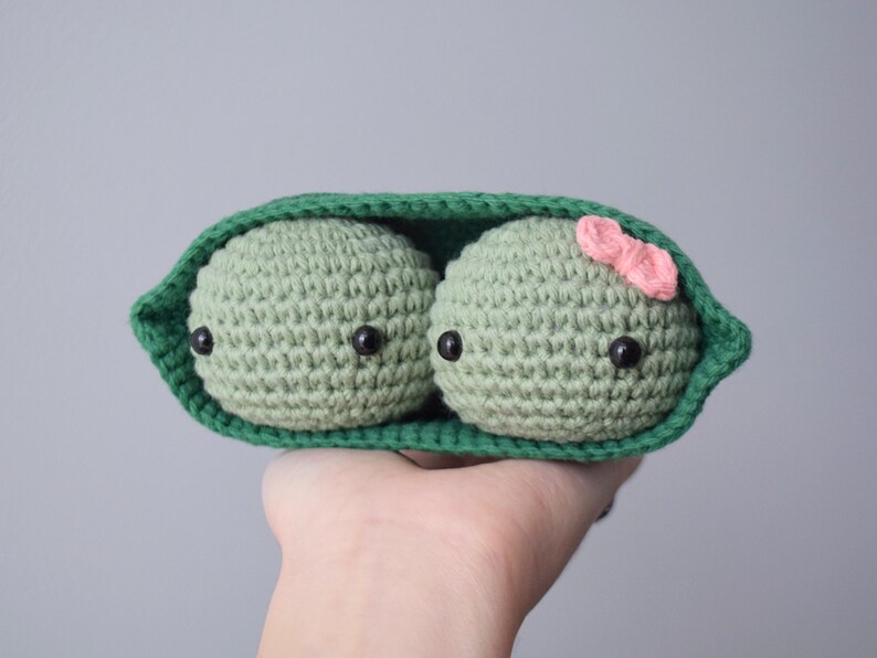 Pu&ograve; includere: Due piselli all'uncinetto in un baccello verde, uno con un fiocco rosa. I piselli hanno occhi neri e sono fatti con filato verde.