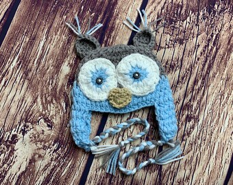 May include: A blue and gray crocheted owl hat with white eyes and a brown nose. The hat has two long ties with tassels.