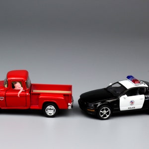 Puede incluir: Una camioneta roja y un coche de policía blanco y negro, ambos modelos de juguete, se enfrentan en un fondo gris.