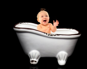 Puede incluir: Una bañera de porcelana blanca con detalles marrones, con un bebé sentado dentro y sonriendo.