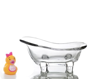 Puede incluir: Una bañera de cristal transparente con cuatro patas, diseñada para contener objetos pequeños. Un pato de goma amarillo con un lazo rosa y la palabra "BABY" en su babero está en primer plano.