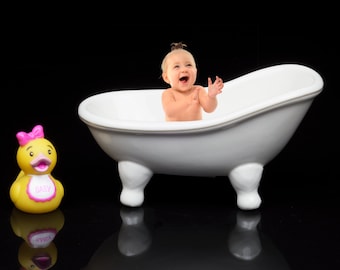 Puede incluir: Una bañera de porcelana blanca con un bebé sentado dentro, sonriendo y aplaudiendo. Un pato de goma amarillo con un lazo rosa y la palabra "BABY" en su pecho está en primer plano.