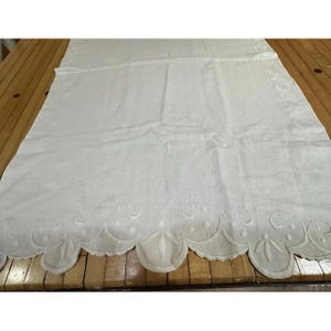 May include: White linen tablecloth with an embroidered scalloped edge. The tablecloth has a delicate floral pattern and an embroidered initial "J" in the center. The scalloped edge features a repeating pattern of circles and stylized leaves.
