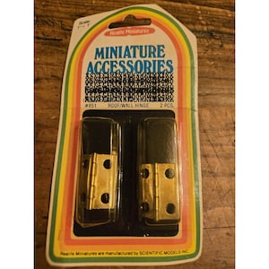 May include: Package of two miniature roof/wall hinges. The hinges have a gold finish and black caps. The packaging is a colorful card with the text "MINIATURE ACCESSORIES" and "Realife Miniatures".
