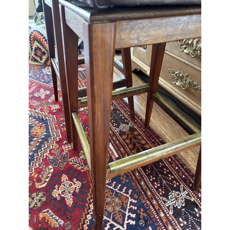 May include: Close-up of two wooden bar stools with brass footrests. The stools have a dark brown finish and a square seat. The background includes a patterned rug with red, blue, and cream colors.