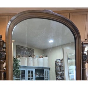 May include: A vintage, arched-top vanity mirror with a wooden frame. The mirror reflects a room with a light fixture, cabinets, and decorative items. The frame has a detailed, carved design.