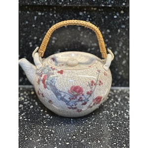 May include: A ceramic teapot with a crackle glaze finish. The teapot features a floral design with red and purple accents. It has a woven handle and a matching lid with a small knob. The spout and handle are white.