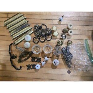May include: A collection of vintage cabinet hardware, including brass pulls, glass knobs, and decorative metal accents. The assortment features various shapes, sizes, and finishes, with a focus on antique and ornate designs. The hardware is arranged on a wooden surface.