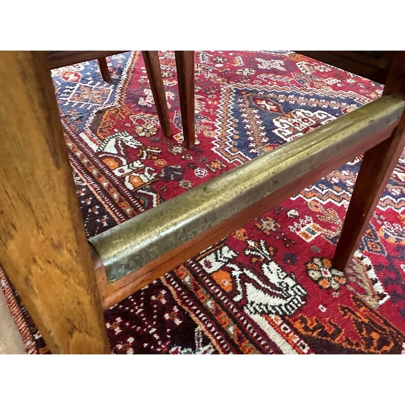May include: Wooden chair with a brass footrest. The chair has a light brown finish and is set against a red and blue patterned rug. The rug features a detailed design with white horse figures.