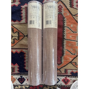 May include: Two rolls of York Wallcoverings wallpaper. The rolls are wrapped in clear plastic and have a light brown, textured appearance. The labels on the rolls include the brand name, product details, and a barcode. The wallpaper is unpasted and scrubbable.
