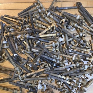 May include: A pile of antique metal hooks and bolts. The hooks have a decorative design and are attached to long, threaded bolts. The metal has a weathered appearance with shades of gold, silver, and black, suggesting age and use.