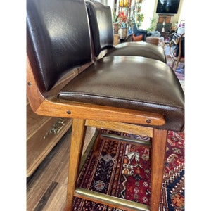 May include: Set of two vintage bar stools. The stools feature dark brown leather seats and backs, with a wooden frame and legs. The legs are connected by a brass footrest. The stools have a mid-century modern design.