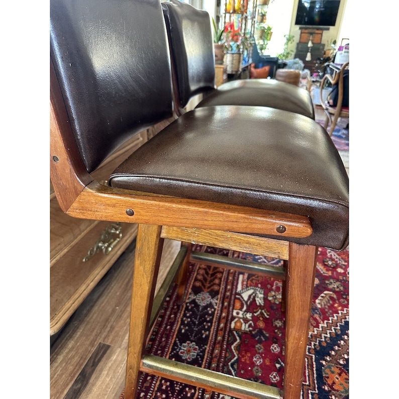 May include: Set of two vintage bar stools. The stools feature dark brown leather seats and backs, with a wooden frame and legs. The legs are connected by a brass footrest. The stools have a mid-century modern design.