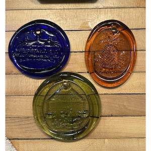 May include: Three decorative glass ornaments in blue, orange, and green. The blue ornament features a building design, the orange one a pineapple, and the green one a barn scene. Each has a hole for hanging.