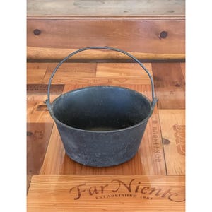 May include: A dark gray cast iron cauldron with a curved metal handle. The cauldron is round with a wide opening and sits on a wooden surface. The handle arches over the top for carrying.