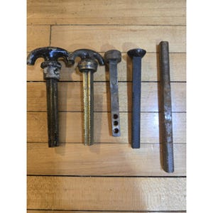 May include: A collection of five antique metal hardware pieces, including threaded rods and handles. The pieces vary in size, shape, and color, with some showing signs of age and wear. The hardware is arranged on a wooden surface.
