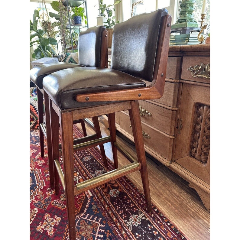 May include: Two vintage bar stools with dark brown leather seats and backs. The stools have wooden frames with brass footrests and armrests. The design is mid-century modern, with a focus on clean lines and natural materials.