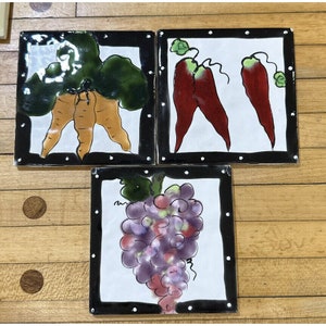 May include: Three square ceramic tiles with black borders and white polka dots. Each tile features a different fruit or vegetable: a bunch of carrots, two red chili peppers, and a bunch of purple grapes.