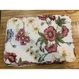 May include: A floral quilted pillow with a scalloped edge. The pillow features a white background with a pattern of large, burgundy, yellow, and blue flowers with green leaves. The pillow is rectangular.