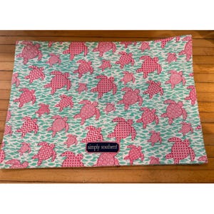 May include: A rectangular, patterned fabric featuring pink sea turtles in various sizes against a teal water background. Some turtles have geometric designs. The fabric has the text "simply southern" on a navy blue label.
