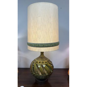 May include: A vintage table lamp with a cream-colored lampshade and a decorative green band. The base is a textured, spherical design in shades of gold and green. The lamp is a decorative lighting fixture.