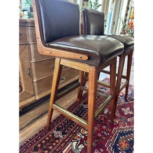 May include: Two vintage bar stools with dark brown leather seats and backs. The stools have wooden frames with brass footrests. The wood has a warm, natural finish. The stools are set on a patterned rug.