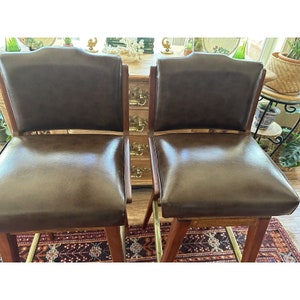 May include: Two brown leather bar stools with wooden frames and brass footrests. The stools have a classic design with curved backs and padded seats. The stools are placed in front of a wooden cabinet.