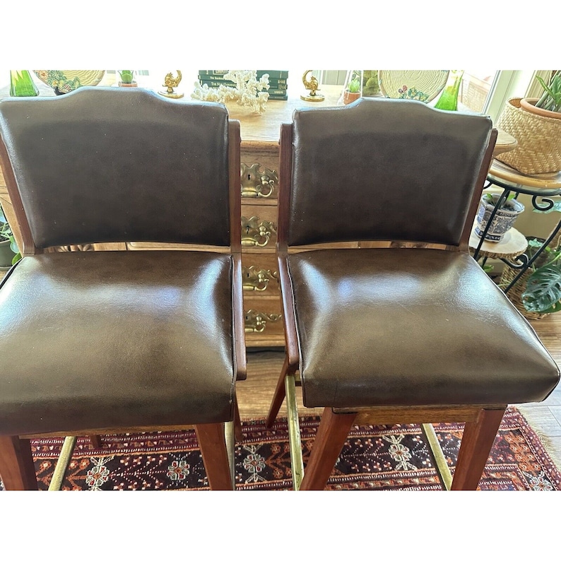 May include: Two brown leather bar stools with wooden frames and brass footrests. The stools have a classic design with curved backs and padded seats. The stools are placed in front of a wooden cabinet.