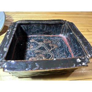 May include: A square ceramic planter with a dark blue glaze and a raised design of a tree in the bottom. The planter has a red rim.