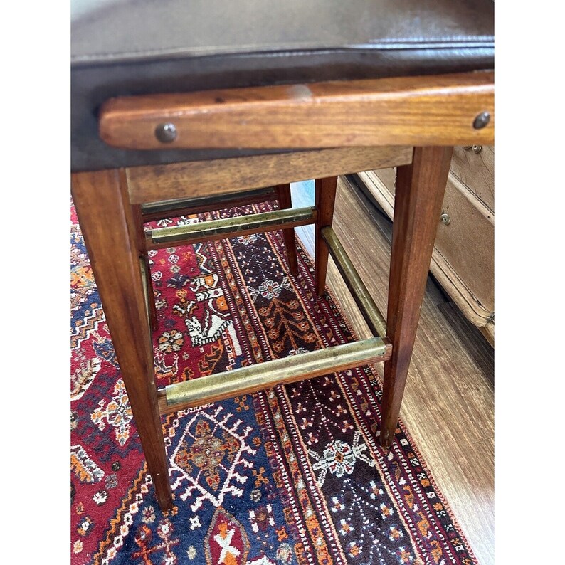 May include: A close-up view of a wooden bar stool with a dark brown leather seat. The stool has a rectangular wooden frame with brass footrests and is set on a patterned rug.
