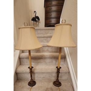 May include: Two matching gold candlestick lamps with beige lampshades. The lamps are on a set of stairs with a wooden dresser in the background.